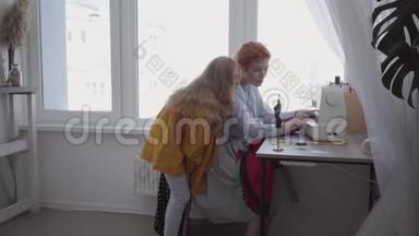 漂亮的红发妈妈把坐在靠窗的桌子旁的衣服缝好，告诉她的女儿如何制作